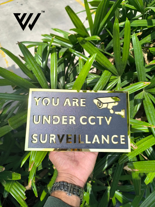 Hand holding a sign with 'You are under CCTV surveillance' text against a green leafy background